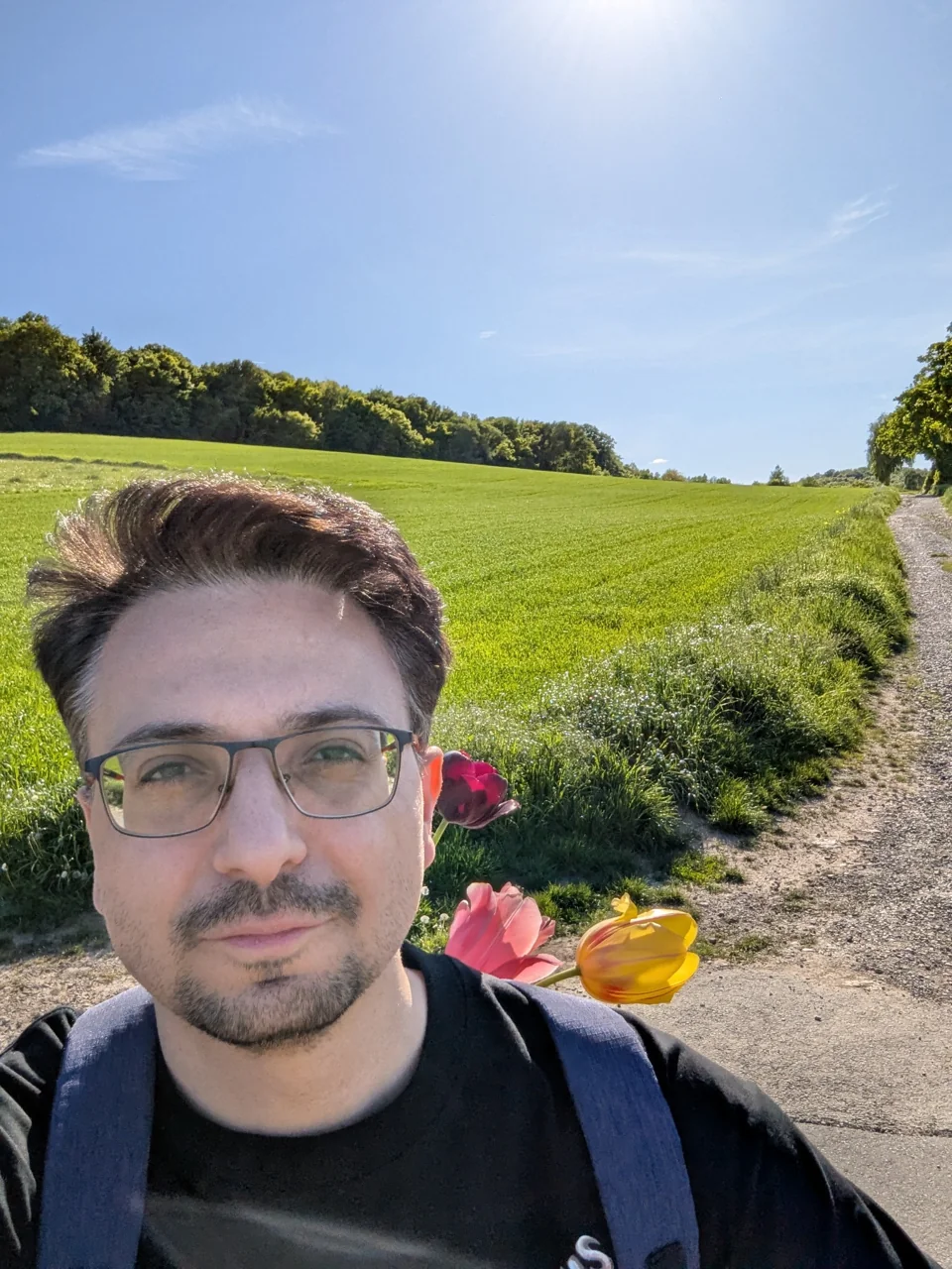 Serdar Kar mit Blumen im Rucksack auf einem Feldweg vor grünen Wiesen und blauem Himmel.